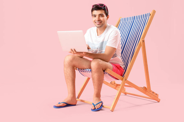 Hombre joven con portátil sentado en la silla de cubierta sobre fondo rosa - Foto, Imagen