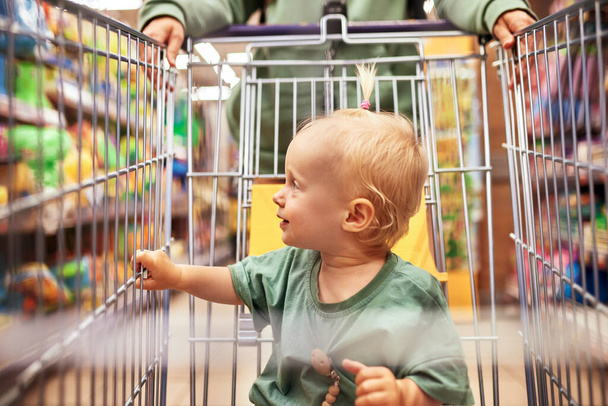 Close-up of cute baby in shopping cart being driven by her mom in supermarket. Mother and little blonde daughter in similar green clothes shopping and having fun. Family weekend, happy childhood - Photo, Image