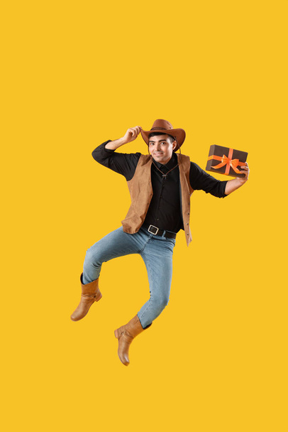 Młody człowiek ubrany na Halloween jako kowboj z prezentem skacząc na żółtym tle - Zdjęcie, obraz