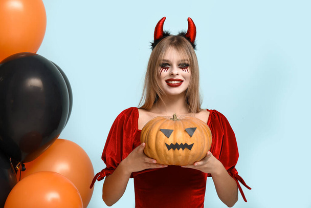 Young woman dressed for Halloween as devil with pumpkin and balloons on blue background - Photo, Image