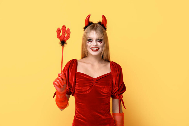 Mujer joven vestida para Halloween como diablo con tridente sobre fondo amarillo - Foto, imagen