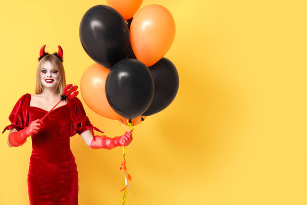 Young woman dressed for Halloween as devil with balloons on yellow background - Фото, изображение