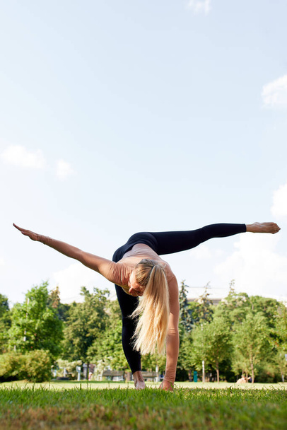 Mujer joven practica yoga en el parque de verano. Chica rubia caucásica delgada en traje negro haciendo ejercicios para fortalecer su cuerpo y espíritu. Gimnasia matutina al aire libre. Estilo de vida saludable - Foto, imagen