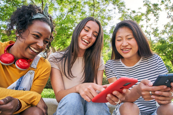 Drei glückliche Frauen mit dem Handy im Freien. Gruppenweise lächelnde College-Freunde checken soziale Netzwerke auf dem Smartphone. Die Klassenkameraden haben Spaß dabei, etwas auf dem Handy anzuschauen. Junge Mädchen flirten online.  - Foto, Bild
