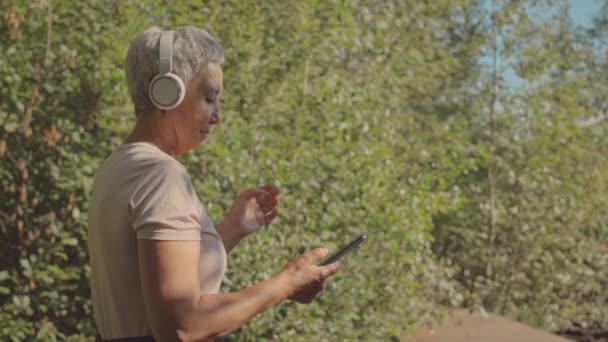 Medium shot of active mature Asian woman in wireless headphones turning on music on smartphone while jogging in forest alone at daytime - Footage, Video