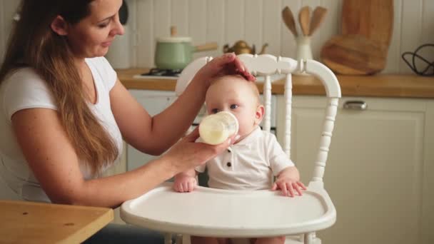 Evde mutlu bir aile. Anne, küçük bebeğini mutfaktaki şişeden besliyor. Yeni doğmuş kız süt içiyor. Bebek emziriyor, süt yiyor ve sandalyeye oturuyor. Bebeği emziren anne. - Video, Çekim