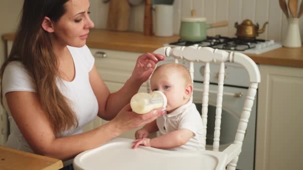 Счастливая семья дома. Мать кормит малыша из бутылочки на кухне. Новорожденная пьет молоко. Младенец сосет молоко сидя на кормящем стуле. Мама грудного младенца - Кадры, видео