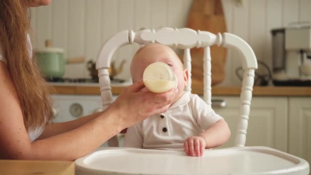 Família feliz em casa. Mãe alimentando criança pequena criança de garrafa na cozinha. Rapariga recém-nascida a beber leite. Bebê infantil chupando comer leite sentado na cadeira de alimentação. Mãe do bebê amamentando - Filmagem, Vídeo