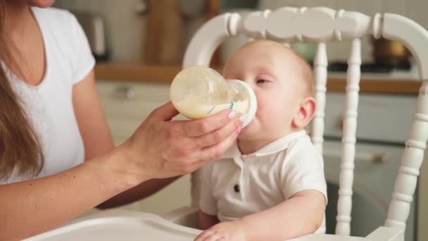 Familia feliz en casa. Madre alimentando a un niño pequeño del biberón en la cocina. Recién nacida bebiendo leche. Bebé lactante chupando comiendo leche sentado en la silla de alimentación. Mamá del bebé lactante - Imágenes, Vídeo