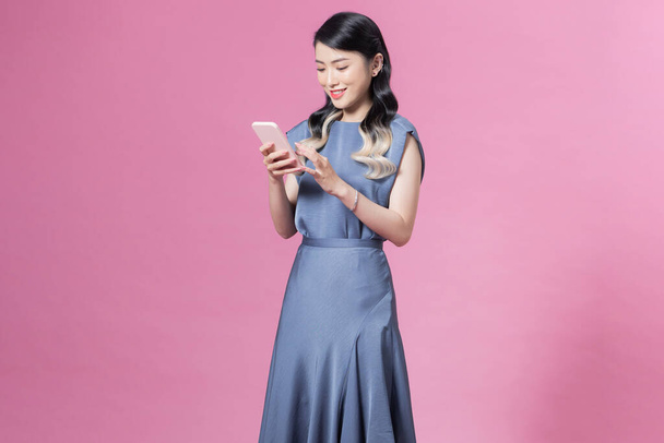 junge Frau trägt blaues Seidenkleid mit Smartphone vor rosa Hintergrund - Foto, Bild