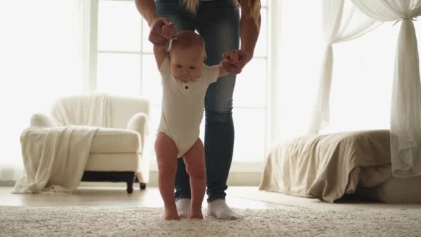 Babyentwicklung. Kleines Mädchen macht erste Schritte zu Hause. Mutter mit Babyhänden hilft Kleinkind bei ersten Schritten im Wohnzimmer. Glückliche Familie zu Hause. Mutter lehrt Baby - Filmmaterial, Video