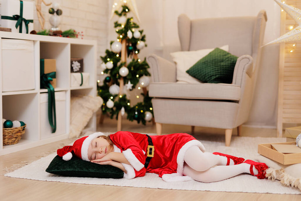 Kırmızı cüce kostümlü küçük kız Noel süslemeli odada uyuyor. - Fotoğraf, Görsel