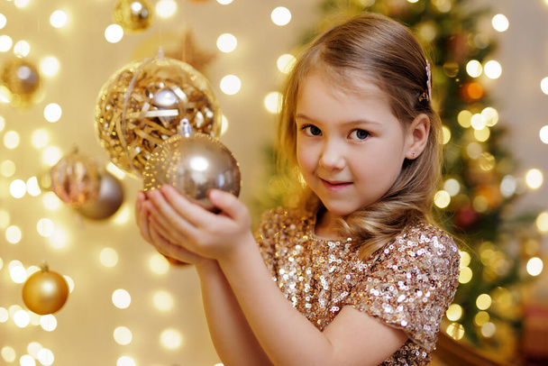Улыбающаяся маленькая девочка держит золотой рождественский бал в украшенной комнате - Фото, изображение