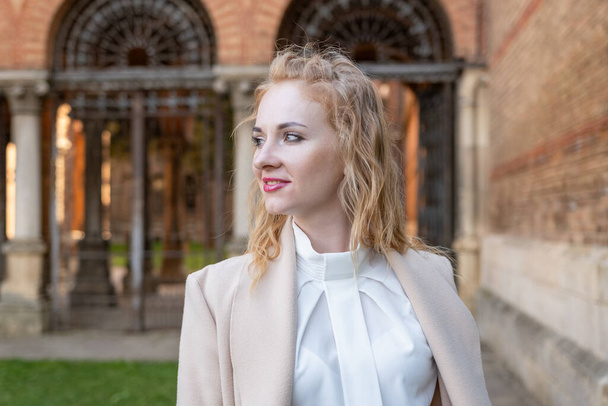 Profile portrait of young woman with blonde curly hair in white blouse and beige coat outside. Girl smiles and looks sideways - Photo, Image