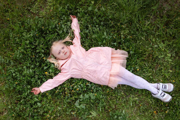 Pembe elbiseli ve ceketli küçük sarışın kız yeşil çimlerin üzerinde uzanıyor ve kolları yanlara doğru uzanıyor. Üst görünüm - Fotoğraf, Görsel