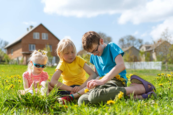Três crianças sentam-se no prado verde com flores e tecem a grinalda de dentes-de-leão. Irmãos relaxar juntos na grama verde - Foto, Imagem