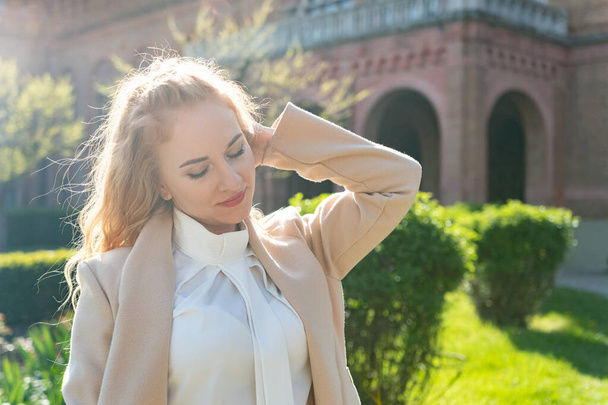 Portrait of stylish business woman in white blouse and beige coat in park enjoying sunny day. Springtime - Photo, Image