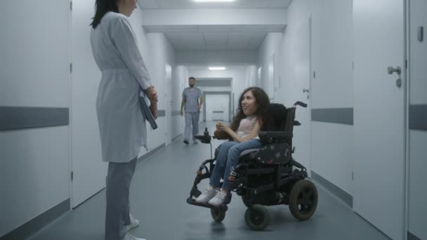Sairaalan käytävä: Nainen, jolla on SMA-vamma pyörätuolissa, keskustelee ammattilaislääkärin kanssa lääkärintarkastuksesta. Naislääkäri konsultoi potilasta nykyaikaisen klinikan tai lääketieteellisen laitoksen valoisassa käytävässä. - Materiaali, video