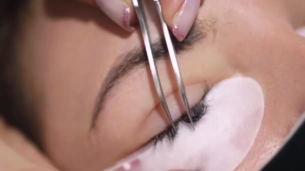 Crop professionele vrouwelijke master met pincet toepassen van kunstmatige wimpers op jonge vrouwelijke client in het gezicht masker in licht moderne salon - Video
