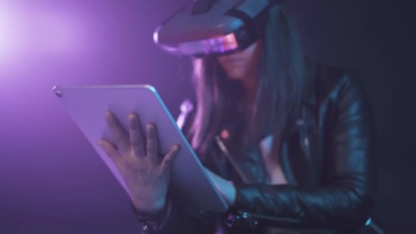 Концентрована анонімна жінка в сучасному гарнітурі VR, що стоїть біля стіни в темній кімнаті з сучасним планшетом в руках під неоновим освітленням
 - Кадри, відео