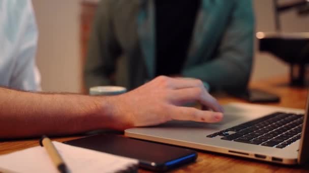 コーヒーショップの木のカウンターに座ってノートパソコンを使いながらノートを取りながら一緒に働くクロップ男性バリスタの側面図 - 映像、動画