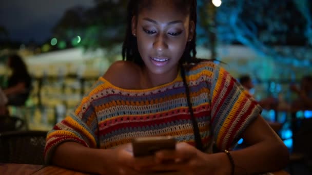 Αφρο-Αμερικανίδα που κάθεται στο street cafe το βράδυ και στέλνει μηνύματα στα social media μέσω smartphone απολαμβάνοντας το καλοκαιρινό Σαββατοκύριακο - Πλάνα, βίντεο