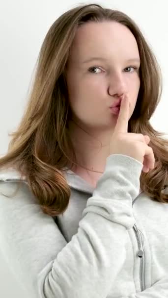 Egy fiatal tinédzser lány az ajkára teszi az ujját, ne mondja el senkinek, hogy mereven forduljon meg, nézzen bele a keretbe, egy ujjal is negatívan. fehér háttér szigorú megjelenés - Felvétel, videó