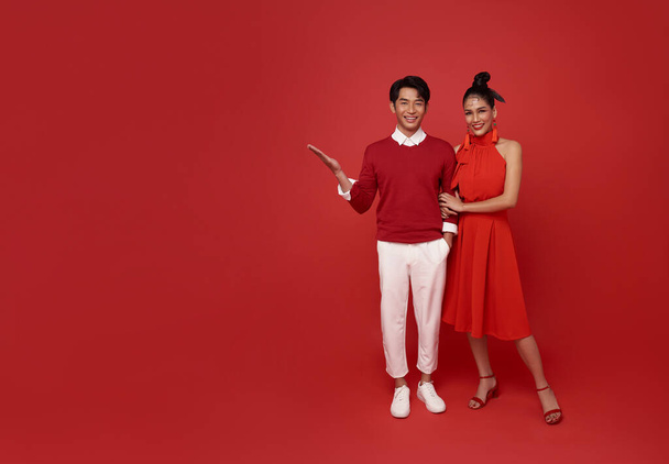 Happy asian couple in red casual attire with open hand present something for blank announcement banner in red background. - Foto, Imagem