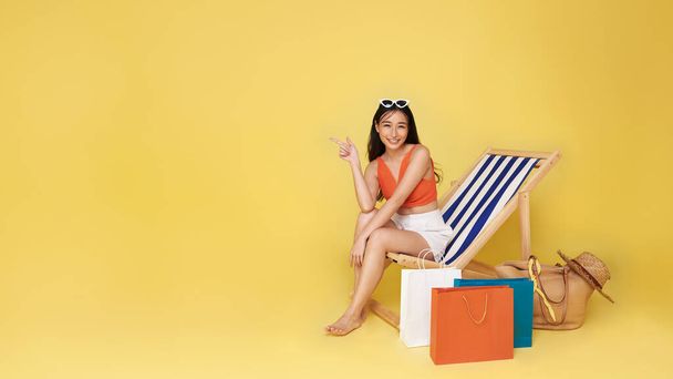 Счастливая азиатская сексуальная женщина в летней одежде с сумкой для покупок сидит на пляжном шезлонге, указывая пальцем на желтом фоне пространства для копирования. Концепция летнего отдыха на пляже. - Фото, изображение