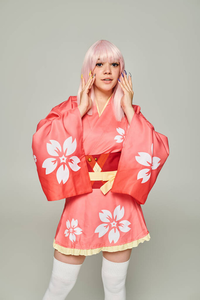 νεαρή ξανθιά γυναίκα με πολύχρωμο μανικιούρ φορώντας ροζ κιμονό και κοιτάζοντας την κάμερα στο γκρι - Φωτογραφία, εικόνα
