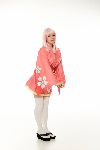 full length of young model with blonde hair posing in pink kimono and white knee socks on white - Photo, Image