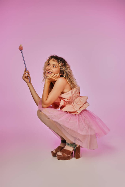 bonita mujer en traje de hada de los dientes con varita mágica en las manos en cuclillas y posando sobre fondo rosa - Foto, Imagen