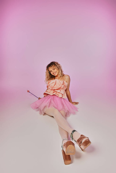 lovely joyous tooth fairy in pink costume sitting on floor with legs crossed on pink backdrop - Photo, Image