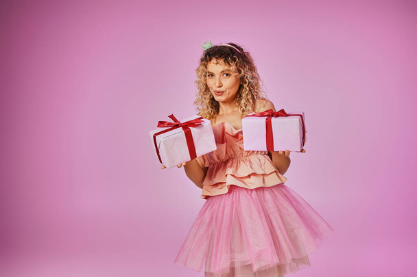 мечтательная блондинка в розовом наряде зубной феи показывает два подарка на камеру на розовом фоне - Фото, изображение