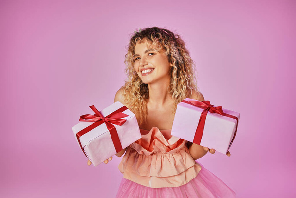 весела кучерява жінка, яка щасливо дивиться на камеру і тримає два подарунки позує на рожевому тлі - Фото, зображення