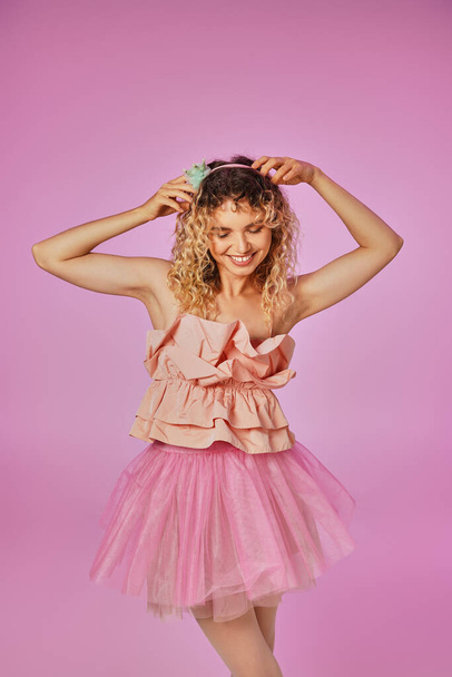 lovely curly haired tooth fairy in pink costume touching her headband and smiling on pink backdrop - Photo, Image