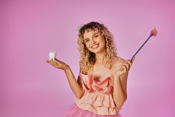 joyous kudrnaté vlasy zubní víla v růžovém kostýmu a čelenka drží baby zub a kouzelnou hůlku - Fotografie, Obrázek