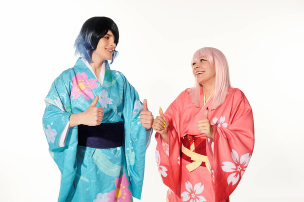 joyful cosplayers in colorful kimonos and paruky showing thumbs up and looking on each other on white - Fotografie, Obrázek