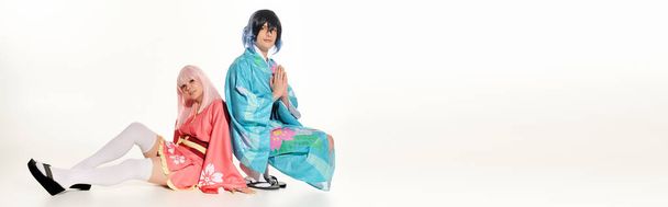 muž v kimonu s modlícími se rukama v blízkosti anime žena v blond paruce sedí na bílém, horizontální prapor - Fotografie, Obrázek