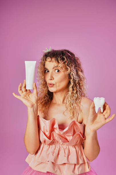 изумленная блондинка в розовом костюме держит молочный зуб и зубную пасту и смотрит в камеру - Фото, изображение