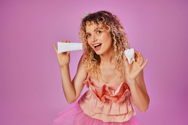 smiling attractive woman in pink costume holding tooth paste and baby tooth on pink backdrop - Photo, Image