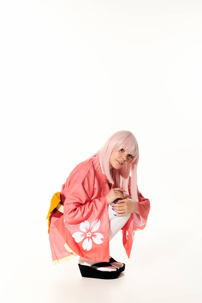 joven mujer de anime en kimono rosa y peluca rubia sentado en las brujas y mirando a la cámara en blanco - Foto, imagen