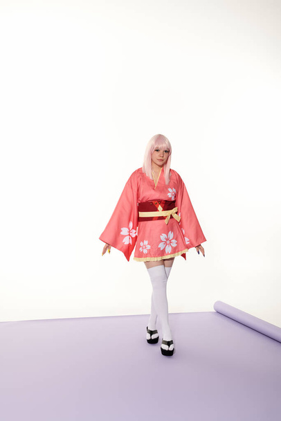 νεαρή γυναίκα anime στυλ σε ροζ κιμονό και ξανθιά περούκα σε μοβ χαλί και λευκό φόντο, cosplay - Φωτογραφία, εικόνα