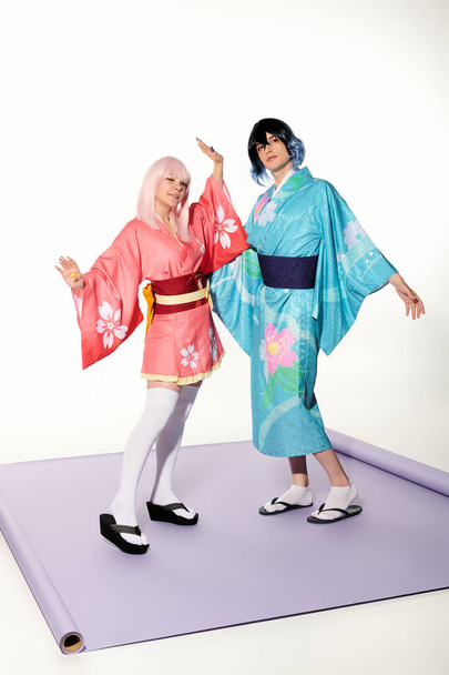jóvenes cosplayers expresivos en coloridos kimonos y pelucas posando sobre alfombra púrpura en estudio blanco - Foto, Imagen