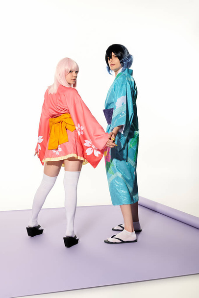 anime estilo pareja en kimonos cogidas de la mano y mirando a la cámara en la alfombra púrpura en estudio blanco - Foto, imagen