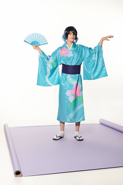 expresivo hombre en kimono azul y peluca posando con abanico de mano sobre alfombra púrpura y fondo blanco - Foto, imagen