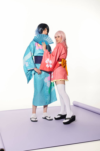 blondynka anime styl kobieta gospodarstwa ręka wentylator w pobliżu mężczyzna w kimono na fioletowy dywan w biały studio - Zdjęcie, obraz