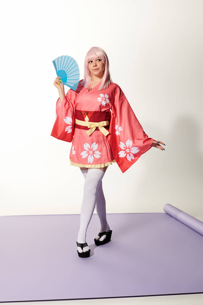 Anime stili kadın pembe kimonolu ve sarı peruklu. Beyaz stüdyoda mor halıda yelpaze. - Fotoğraf, Görsel
