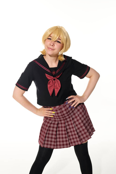 jeune femme ludique en perruque blonde jaune et uniforme scolaire avec les mains sur les hanches sur blanc, cosplay - Photo, image