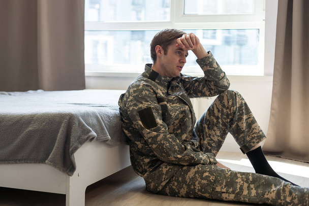 Stresszes veterán katona, tele depressziós gondolatokkal és emlékekkel. Szomorú, depressziós fiatalember álruhás katonában ül a kanapén, és a háborún jár az esze. PTSD és terápiás koncepció - Fotó, kép